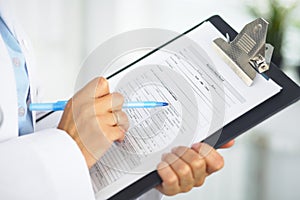 Close-up of a female doctor filling up medical form at clipboard while standing straight in hospital