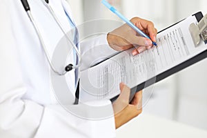 Close-up of a female doctor filling up medical form at clipboard while standing straight in hospital
