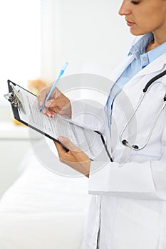 Close-up of a female doctor filling up medical form at clipboard while standing straight in hospital