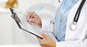 Close-up of a female doctor filling up medical form at clipboard while standing straight in hospital