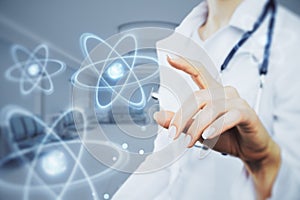 Close up of female doctor hand pointing at abstract glowing atom hologram on blurry hospital or clinic interior background.