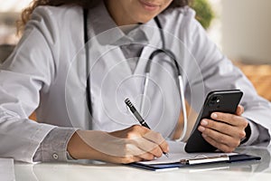 Close up female doctor wearing uniform holding smartphone, taking notes