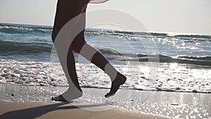 Close up of female feet running on wet sand and sea water at the beach with waves at background. Legs of young woman