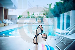 Close up female feet in sandals relaxing near blue water of the swimming pool in hotel in evening time. Calm, slow, harmony