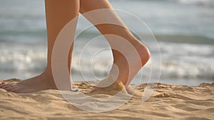 Close up of female feet walking on golden sand at the beach with ocean waves at background. Legs of young woman stepping