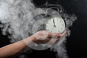 Close-up of a female hand holding a clock on a black background in smoke. Alarm clock at midnight in a mystical fog.