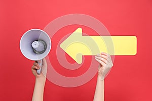 Close up of female hand holding megaphone pointing on big yellow arrow, isolated over red studio background wall with copy space