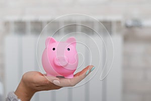 Close up of a female hand holding piggy bank with heating radiator on background