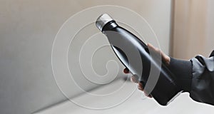 Close-up of female hand holding reusable eco steel thermo water bottle of black on background of office table.