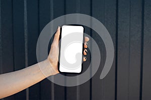 Close-up of female hand holding smartphone with blank on screen, on background of black metal facade of modern house. Mockup