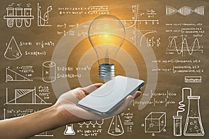 Close up of female hand holding smartphone with glowing light bulb on chalkboard wall background with mathematical formulas.