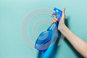 Close-up of female hand holding spray bottle for cleaning with blue pump, on cyan background.