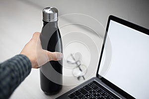 Close-up of female hand holding steel eco thermo bottle for water over office desk, laptop with mockup and glasses.