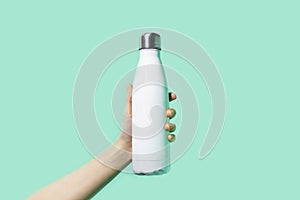 Close-up of female hand, holding white reusable steel stainless eco thermo water bottle on background of cyan, aqua menthe color.