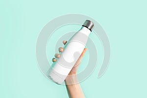 Close-up of female hand, holding white reusable steel stainless eco thermo water bottle with mockup, isolated on cyan color.