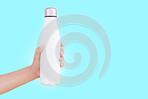 Close-up of female hand holding white reusable steel thermo water bottle isolated on background of cyan color with copy space.