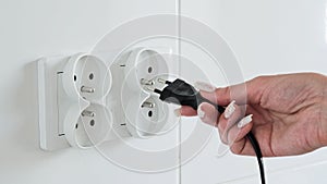Close-up of a female hand inserting a black electric plug into a double European electrical socket  on a white wall.