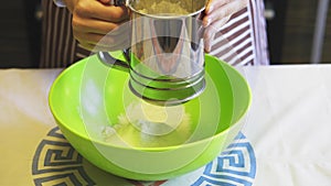 Close-up of a female hand sifting flour with a sieve mug in a green bowl in the home kitchen. Cooking pancakes