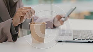 Close-up female hand of sitting businesswoman stirring coffee in disposable cup at table with laptop holding mobile