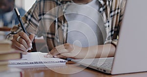 Close-up of female hand writing in notebook and typing with laptop at desk in university