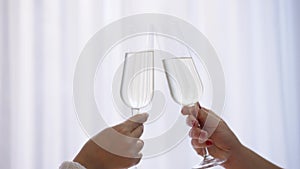 Close-up female hands clinking champagne glasses indoors. Two unrecognizable Caucasian young women toasting booze on