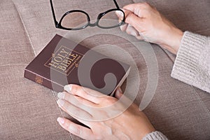Close-up of female hands with glasses and Holy Bible on sofa at home