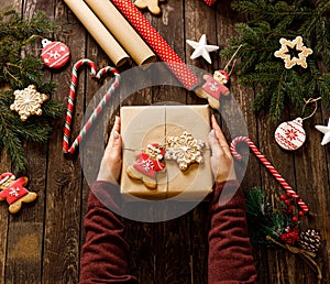 Close up of female hands holding a Christmas gift on a wooden table decorated in winter holiday motives with cookies, biscuits,