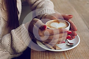 Close-up female hands holding cup with coffee cappuccino with foam with pattern heart. Perfect red gel polish manicure. Wood