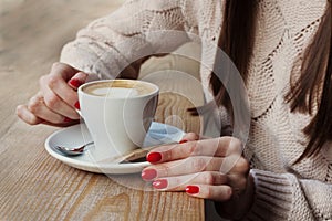 Close-up female hands holding cup with coffee cappuccino with foam with pattern heart. Perfect red gel polish manicure. Wood
