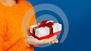 Close-up of female hands holding white gift box with red bow on background of blue color with copy space. Wearing orange sweater.