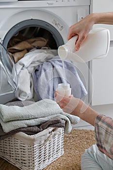 Close up of female hands pouring liquid laundry detergent into cap. Washer machine and clothes with wicker basket in background