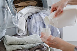 Close up of female hands pouring liquid laundry detergent into cap. Washer machine and clothes with wicker basket in background