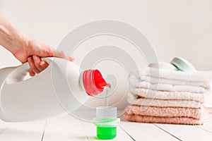 Close up of female hands pouring liquid laundry detergent into cap on white rustic table with towels on background in