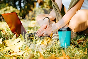 Close-up of female hands typing on laptop keyboard, teenage girl sitting on green grass with device and cup of coffee