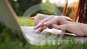 Close-up of female hands typing text on a laptop keyboard in a park at sunset. The concept writer is writing a book in