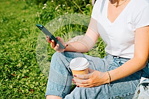 Close-up of female hands using mobile phone and holding disposable cup of coffee outdoors.