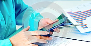 Close-up of female hands using smart phone while working on computer at modern office interior, businesswoman typing