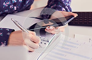 Close-up of female hands using smart phone while working on computer at modern office interior, businesswoman typing