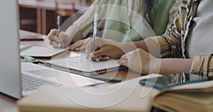 Close-up of female hands witing in notebooks at table with laptop while students learning on campus