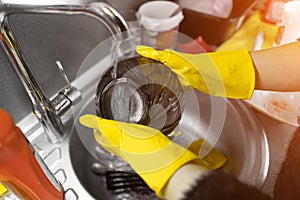 Close up of female hands in yellow protective rubber gloves washing white plate with blue cleaning sponge against kitchen sink