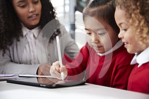 Close up of female infant school teacher sitting at a desk in a classroom helping two schoolgirls wearing school uniforms using a photo