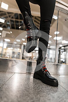 Close up of female legs in black jeans in fashionable black leather boots with red shoelaces in the mall. Stylish young woman