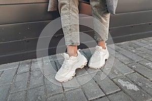 Close-up female legs in fashion jeans in stylish leather sneakers near building in city. Fashionable woman in casual wear on walk