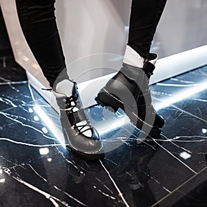 Close up of female legs in trendy leather black-white boots in a modern room. Stylish girl in jeans in fashionable shoes stands in
