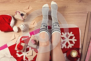 Close-up of female legs in warm socks with a deer, christmas presents, wrapping paper, decoration and cup of cappuccino top view.