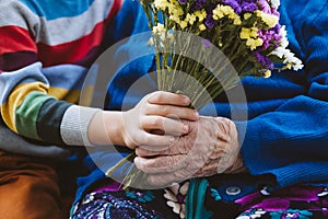 Close up female senior hand holding kid hand and flowers bouquet. Preschooler boy supporting his great-grandmother