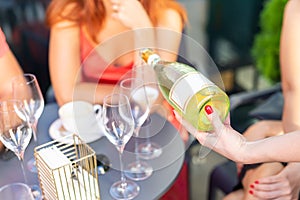 Close-up female waiter holding bottle of white champagne or prosecco at outdoor party on bright summer day. Girls hen