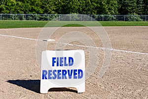 Close up of Field Reserved sign on empty local baseball field, third base and baseline, sunny day with woods in the background