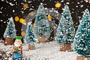 Close-up of figure of the Magus King Melchior with artificial Christmas trees, snow falling, on wooden table with snow, selective