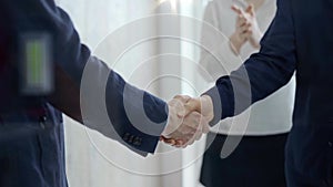 Close-up of a firm handshake between two professionals with a colleague applauding in the background.